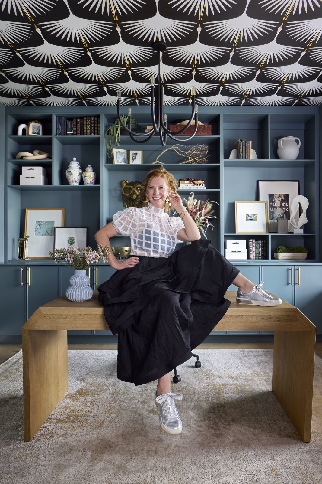 Kendra Stewart, founder of E+E Design Consulting, Denver, in a black skirt and sheer white top, seated on a wood desk beneath a bold crane-patterned ceiling.