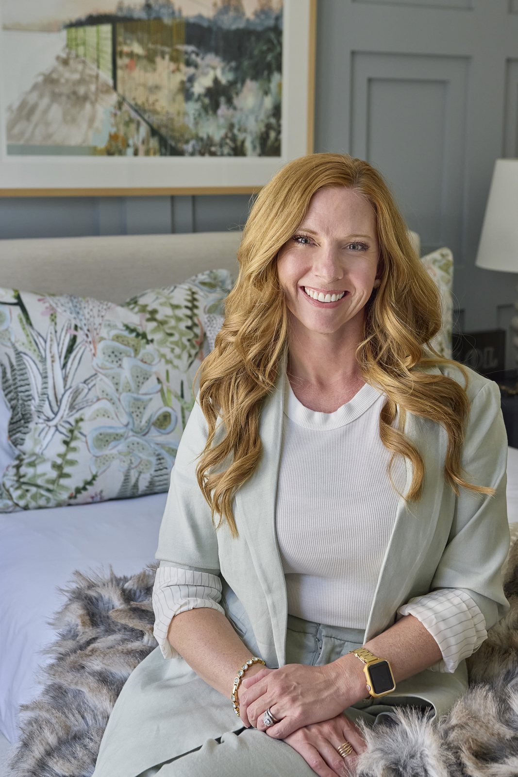 Kendra Stewart, founder of E+E Design Consulting in Denver, smiling warmly while seated on a bed in a soft blue-paneled bedroom.