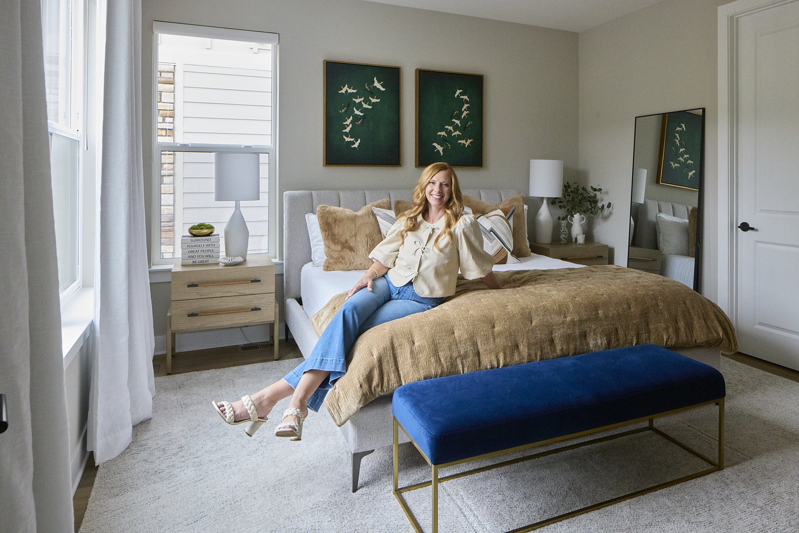 Kendra Stewart, founder of E+E Design Consulting, Denver, seated on a bed in the finished Canyons project bedroom in Castle Pines, with green crane artwork above the headboard.