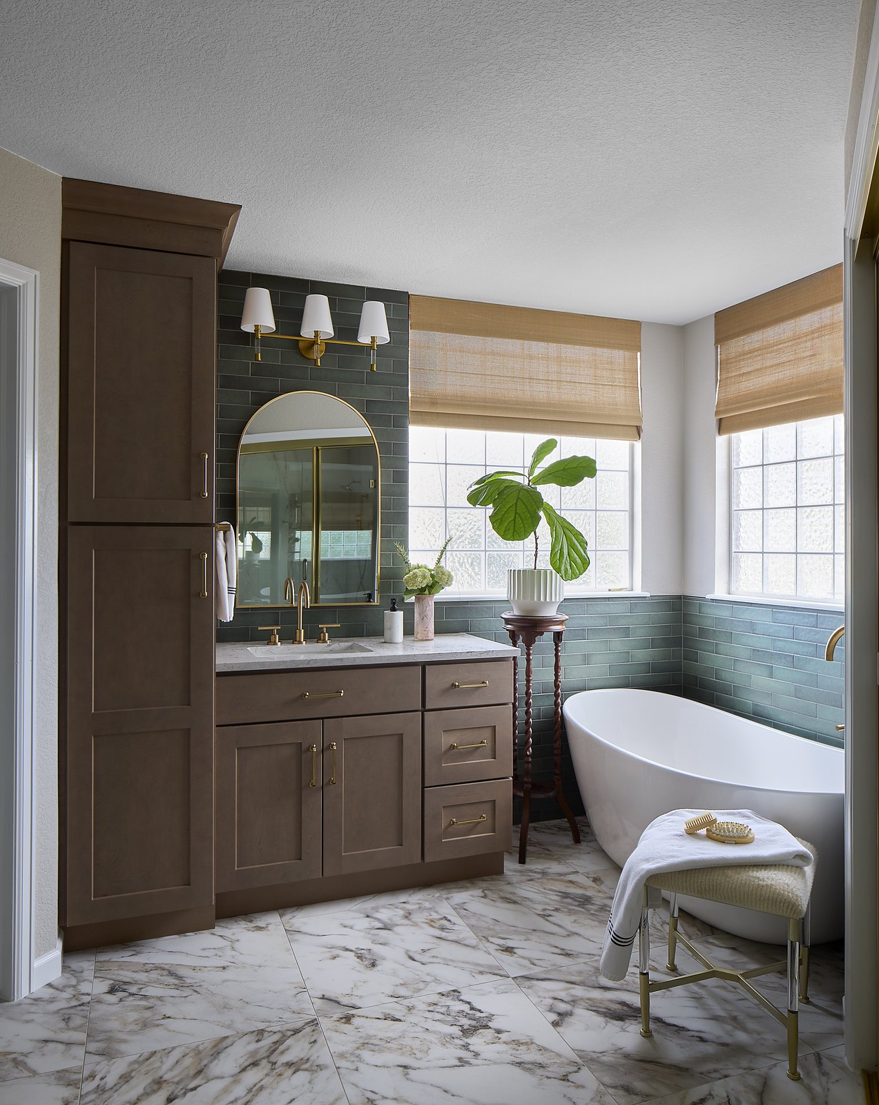 Primary bathroom with a freestanding soaking tub, green tile wainscot, warm wood vanity, arched brass mirror, and woven shades in Canterberry Crossing, Parker