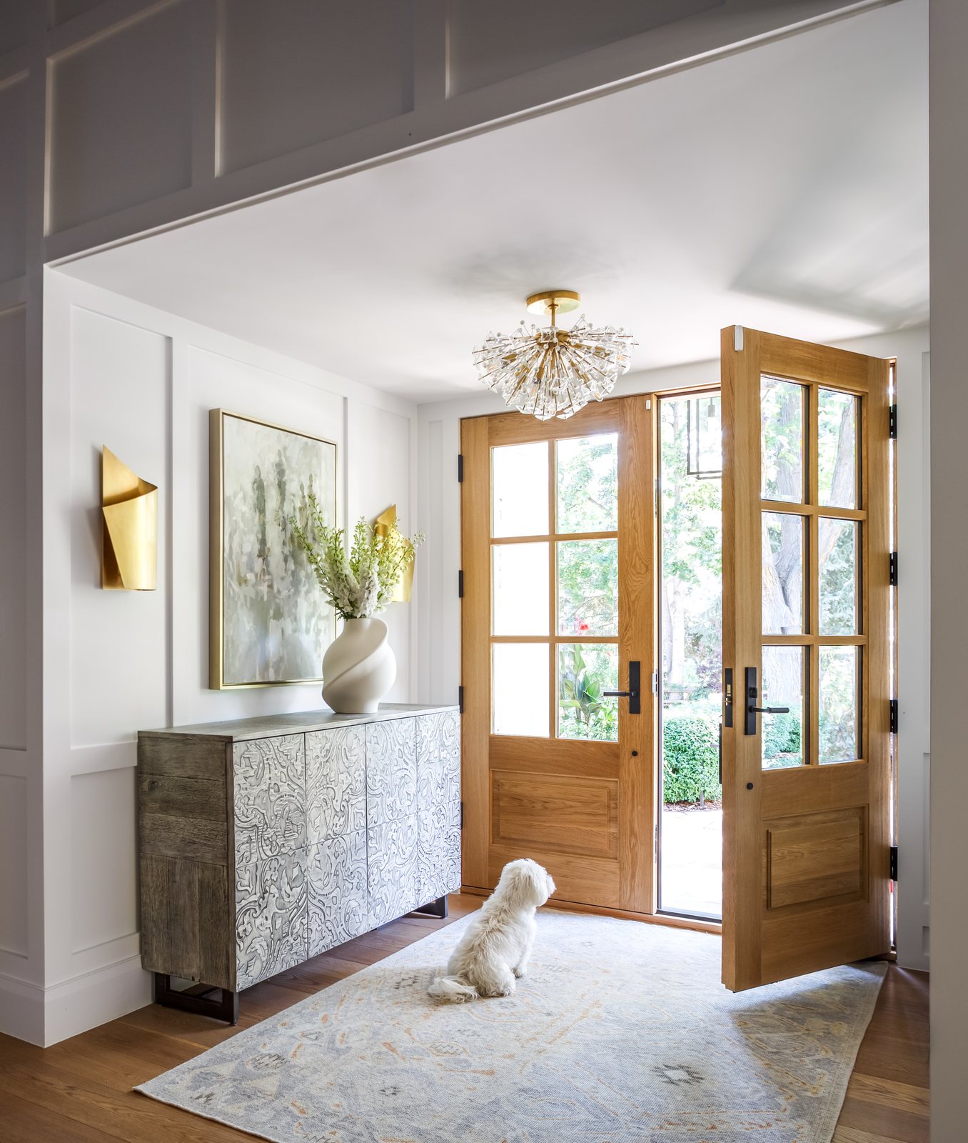 A small white dog waits by open white-oak front doors in the foyer of the Greenwood Village whole-home remodel.