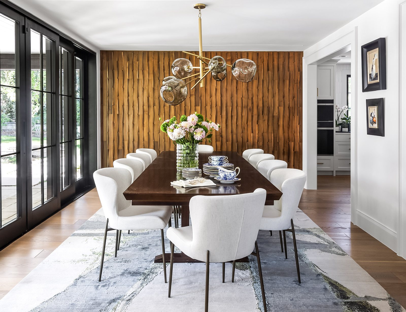 Dining room with a sculpted wood accent wall, brass globe chandelier and white curved chairs in the Greenwood Village whole-home remodel.