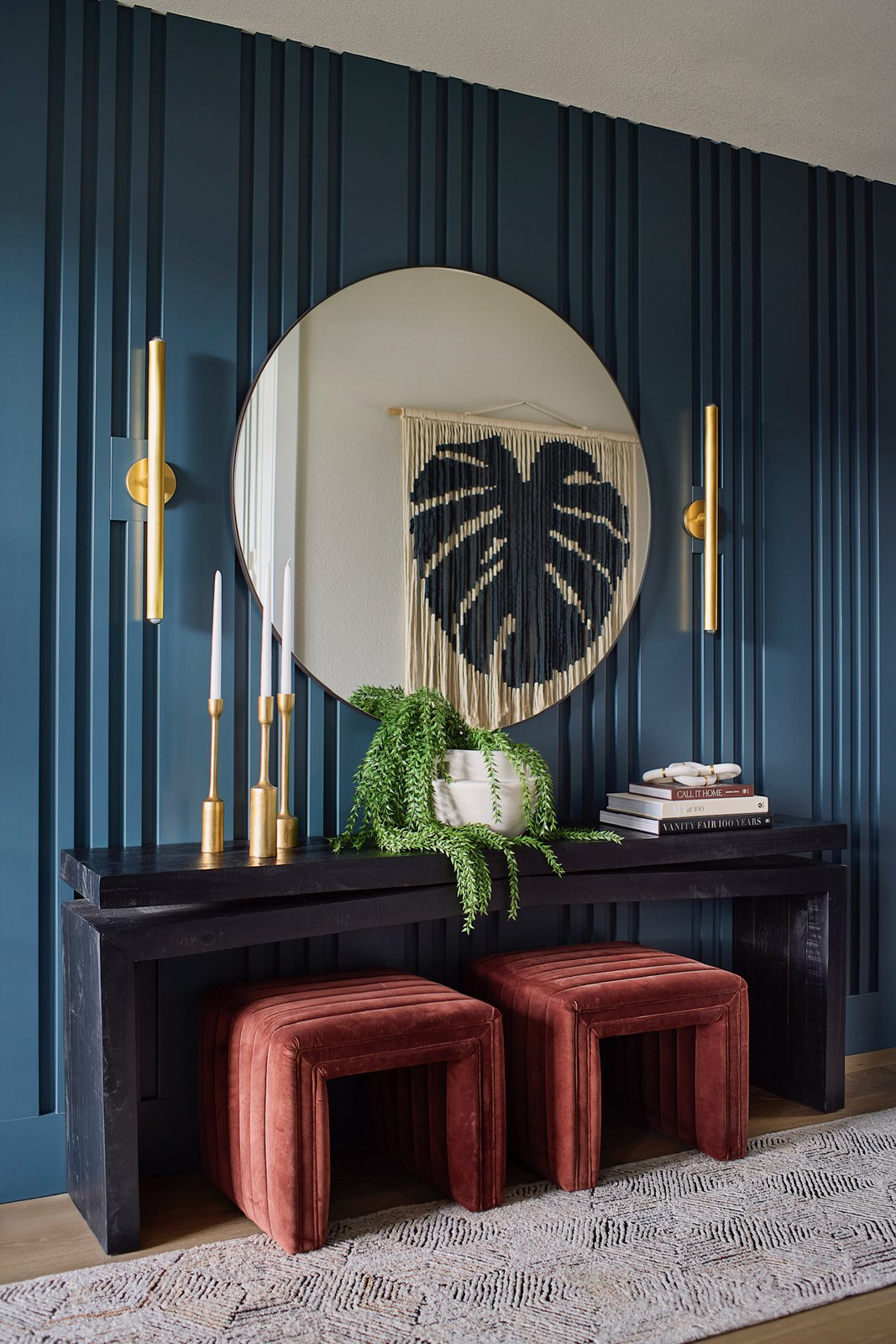 Round mirror, brass sconces and rust velvet ottomans against a navy slat-panel wall at the entry console in The Canyons new build, Castle Pines.