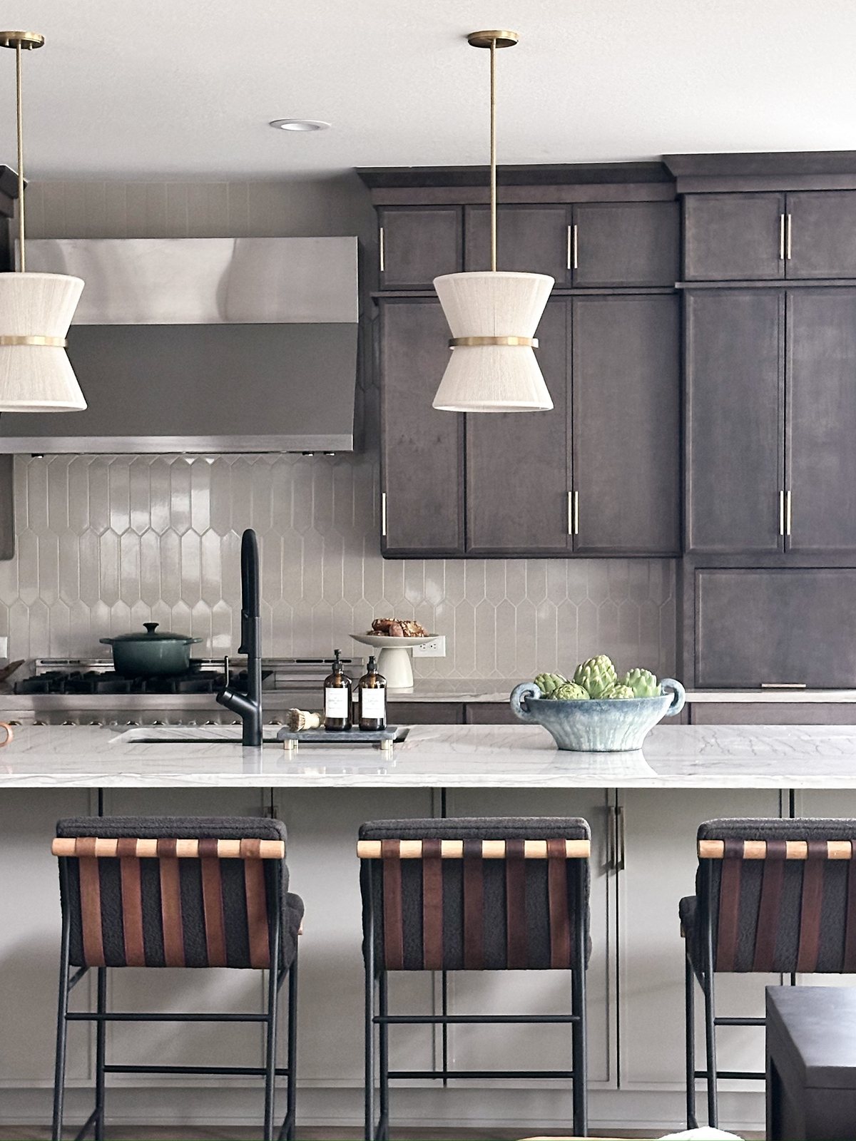 Range wall with stainless hood, picket-tile backsplash and hourglass pendants over the marble island in The Canyons kitchen, Castle Pines.