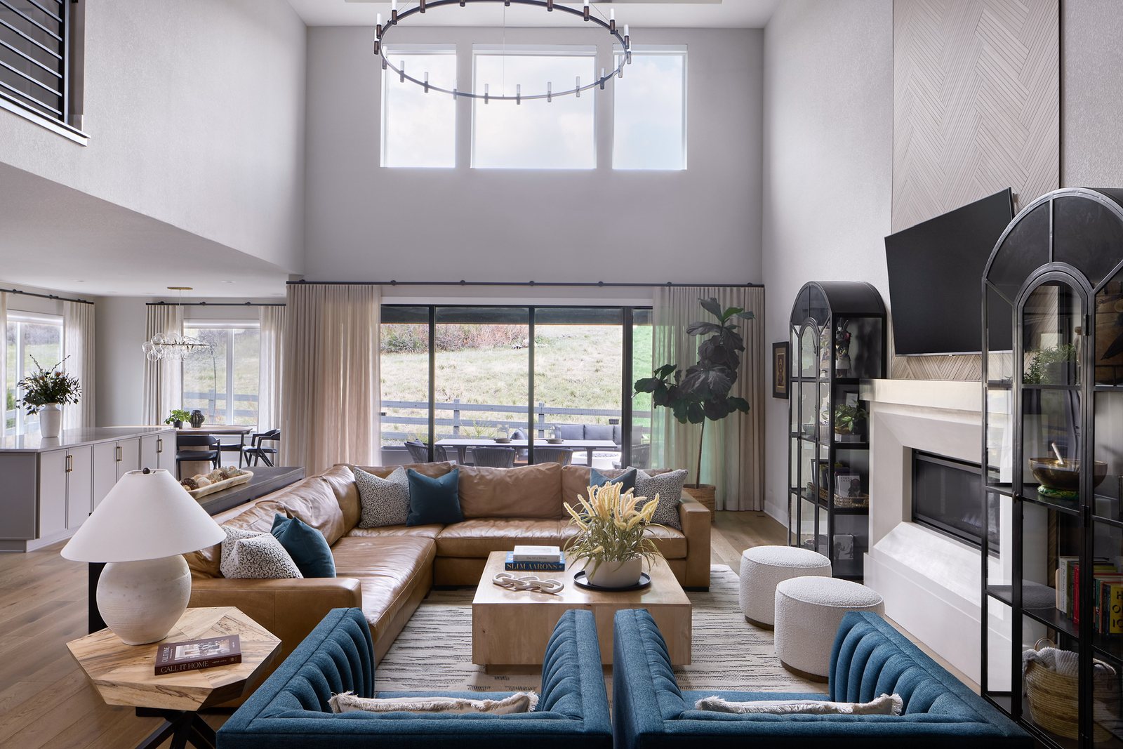 Two-story great room with a tan leather sectional, blue armchairs and ring chandelier opening to the kitchen in The Canyons semi-custom new build, Castle Pines.