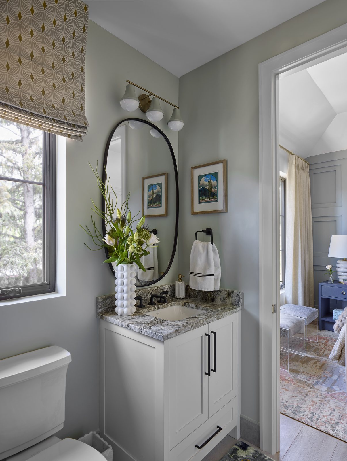 The adjoining bathroom in the Washington Park showhouse in Denver, with a granite-topped white vanity, black oval mirror, and a view through to the bedroom.