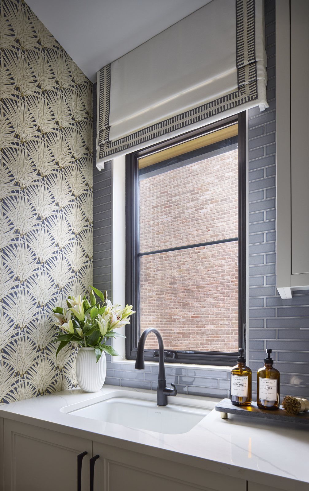 Laundry sink detail in the Washington Park showhouse in Denver, with a black faucet, blue glass tile, a trimmed roman shade, and lilies on the counter.