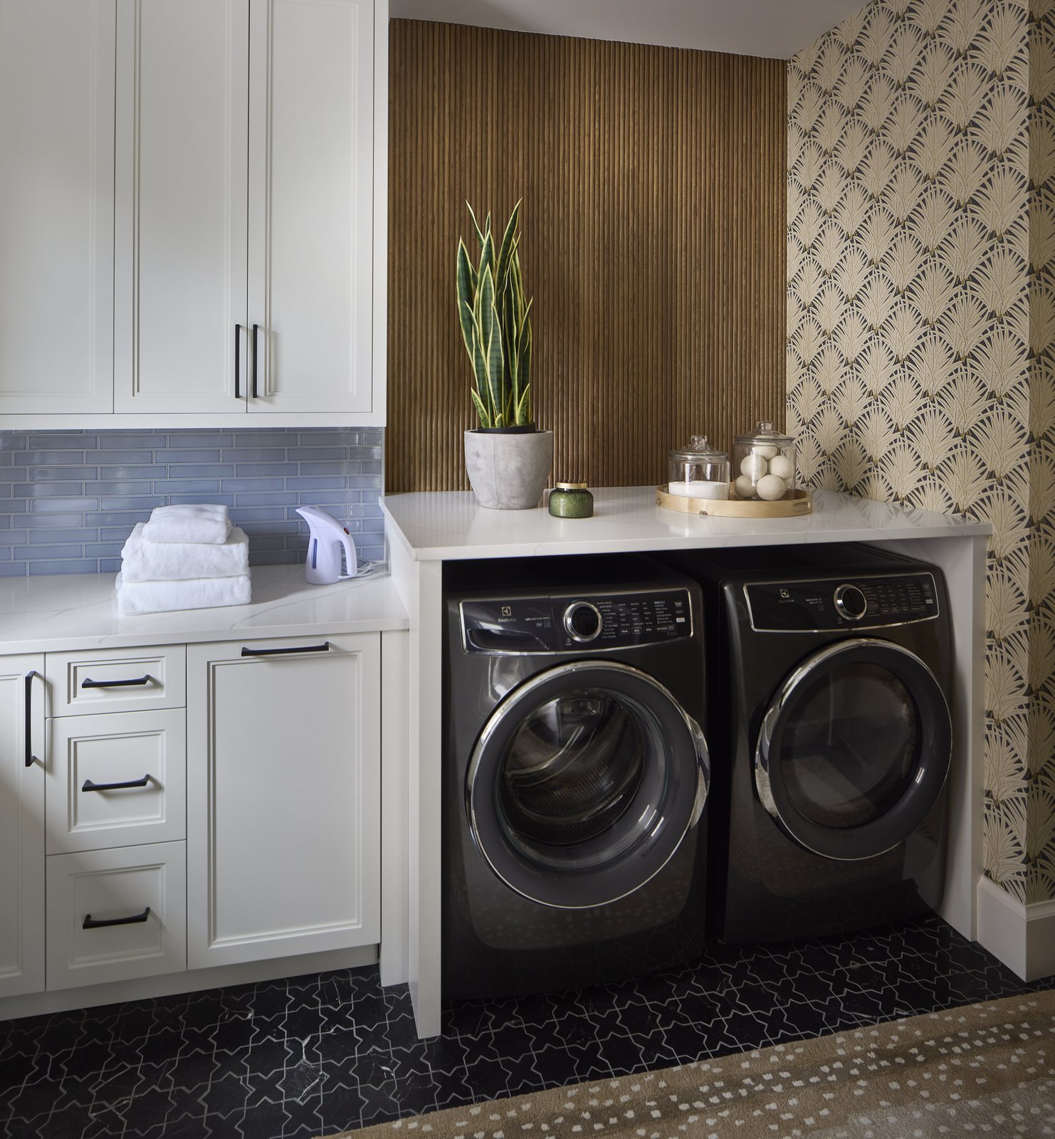Black Electrolux washer and dryer under a quartz counter in the Washington Park showhouse laundry room in Denver, framed by fan-motif wallpaper and a wood slat wall.