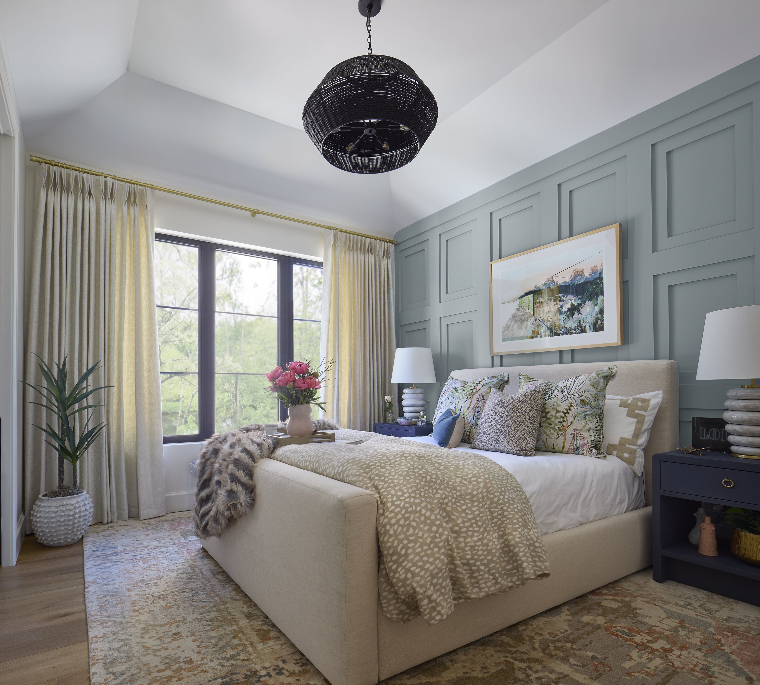 Wide view of the vaulted bedroom in the Washington Park showhouse in Denver, with a blue-gray paneled accent wall, black woven pendant, and a bed dressed in botanical pillows.